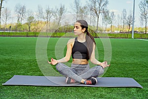 Young happy and beautiful brunette girl doing yoga workout in park, sitting in lotus pose meditating enjoying retreat in balance a