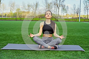 Young happy and beautiful brunette girl doing yoga workout in park, sitting in lotus pose meditating enjoying retreat in balance a
