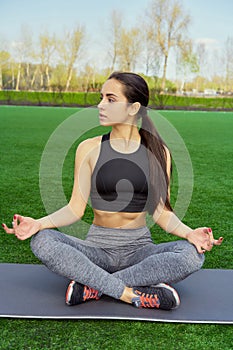 Young happy and beautiful brunette girl doing yoga workout in park, sitting in lotus pose meditating enjoying retreat in balance a