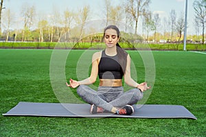 Young happy and beautiful brunette girl doing yoga workout in park, sitting in lotus pose meditating enjoying retreat in balance a