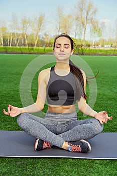 Young happy and beautiful brunette girl doing yoga workout in park, sitting in lotus pose meditating enjoying retreat in balance a