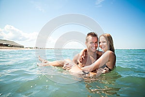 Young happy beautiful couple woman and man standing in blue still sea water and smiling with sandy beach at background