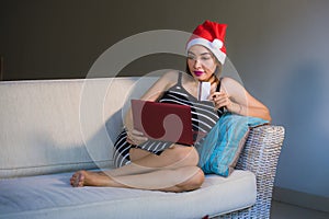 Young happy and beautiful girl relaxed at home couch in Santa hat using laptop computer paying for Christmas present with credit c