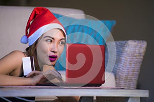 Young happy and beautiful girl relaxed at home couch in Santa hat using laptop computer paying for Christmas present with credit c