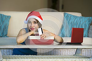 Young happy and beautiful girl relaxed at home couch in Santa hat using laptop computer paying for Christmas present with credit c