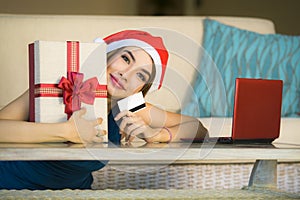 Young happy and beautiful girl relaxed at home couch in Santa hat using laptop computer paying for Christmas present with credit c