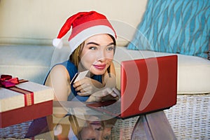 Young happy and beautiful girl relaxed at home couch in Santa hat using laptop computer paying for Christmas present with credit c