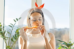 Young happy beautiful woman eating orange slice at home near the window