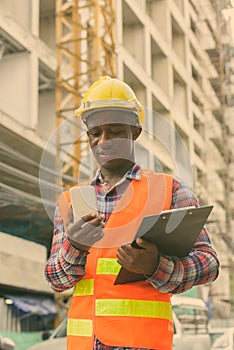 Young happy black African man construction worker smiling while holding clipboard and using mobile phone at building