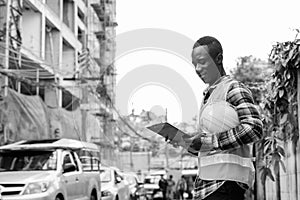 Young happy black African man construction worker smiling while reading on clipboard and holding hard hat at building