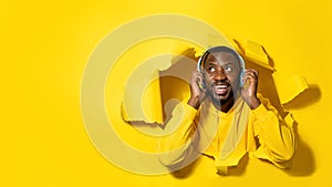 Young happy black man peeking out hole isolated on yellow background and listening to music with headphones