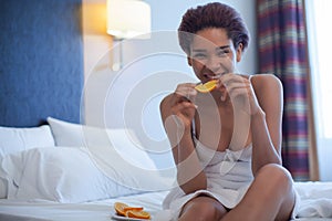 Young happy black woman sits in a bed and eating orange slice.