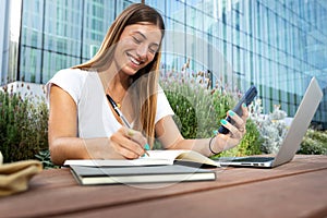 Young happy blonde female college student using phone and laptop to study, taking notes on notebook.