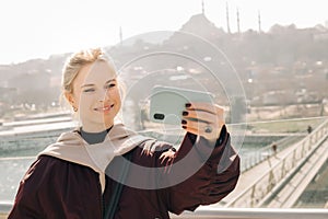 Young happy blonde in jacket makes selfie against background of glass window.