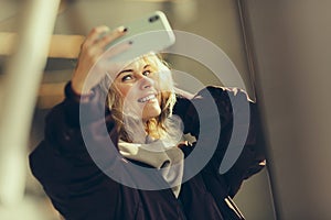 Young happy blonde in jacket makes selfie against background of window