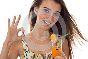 Young happy brunette girl in sarafan with floral pattern drinks orange cocktail smiling on camera and showing OK