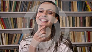 Young happy brunette girl is standing in library, tucking hair, laughing