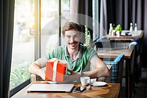 Young happy businessman in green t-shirt sitting and holding big red gift box and looking at camera with toothy smile. business