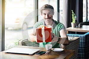 Young happy businessman in green t-shirt sitting and holding and giving big red gift box. looking at camera with toothy smile