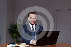 Young happy businessman smiling while using his laptop. Portrait of smiling business man reading message and working in office.