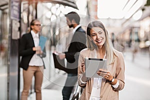 Happy businesswoman holding digital tablet outside of modern building, businesspeople in background