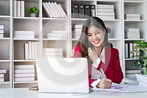 Young happy businesswoman talking on cellphone make notes and using laptop computer working online in modern office.