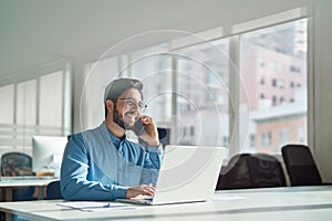 Young happy busy latin businessman talking on cell phone using laptop in office.