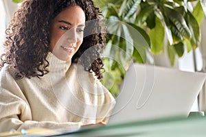 Young happy latin girl student using laptop looking at computer at home office.