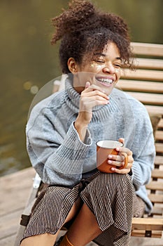 Young happy carefree african american woman with cup of hot tea relaxing near pond in autumn nature