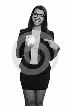 Young happy Caucasian businesswoman smiling and holding piggy bank while pointing finger