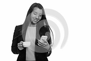 Young happy Caucasian businesswoman smiling while using mobile phone and holding coffee cup