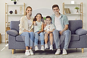 Young happy Caucasian family with two children watching TV together sitting on sofa in living room.