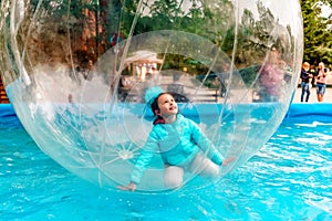 Young happy Caucasian girl playing inside aqua zorb floating on water in city park. Leisure outdoor children attractions and