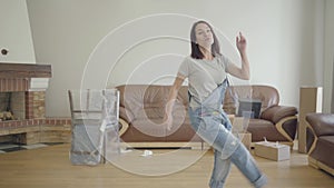 Young happy Caucasian woman dancing in the living room with unpacked furniture and sitting down on the floor. Adult girl