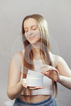 Young happy caucasian woman with long flowing hair is opening unbranded white jar with cosmetics