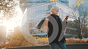 Young happy caucasian woman with shoping bags using phone in the city park