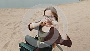 Young happy caucasian woman in the wheelchair on the sandy beach making heart with her hands, overshoulder shot