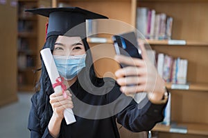 A young happy cheerful Asian woman university graduates in graduation gown and cap wears a face mask holds and shows a degree
