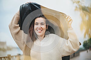 Young happy cheerful woman under rain covering head with coa while standing on city street