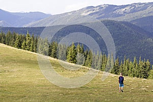 Young happy child boy with backpack walking in mountain grassy valley on background of summer woody mountain. Active lifestyle,