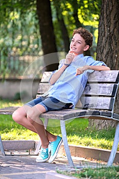 Young happy child boy relaxing sitting on bench in summer park. Positive kid enjoying summertime outdoors. Child