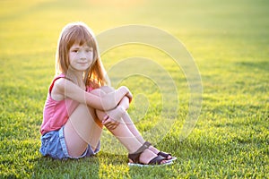 Young happy child girl resting while sitting on green grass lawn on warm summer day