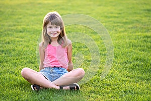 Young happy child girl resting while sitting on green grass lawn on warm summer day