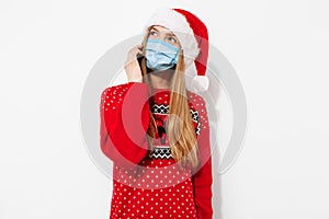 Young happy christmas woman, wearing a Santa Claus hat and a medical mask on her face, talking on the phone, holding a smartphone