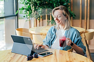 Young happy concentrated woman freelancer working on a laptop in a cafe and drinking berry smoothie.