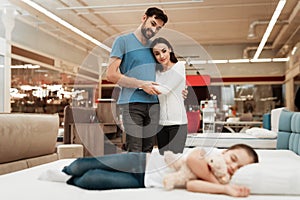 Young happy couple in background enjoying sleeping little girl. Choosing mattress in store.