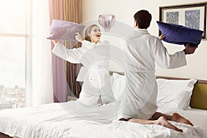 Young happy couple in bathrobe fight pillows on bed