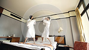 Young happy couple in bathrobe fight pillows and have fun on bed in hotel during their honeymoon vacation