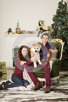 Young happy couple cuddling adorable akita inu dog while sit on stylish retro armchair for Christmas holidays at home. Christmas