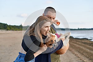 Young happy couple with dog standing on beach. Beautiful girl bitting Corgi puppy ear